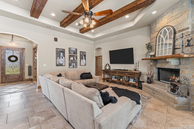 tiled living room featuring a tray ceiling, beam ceiling, a stone fireplace, and ceiling fan