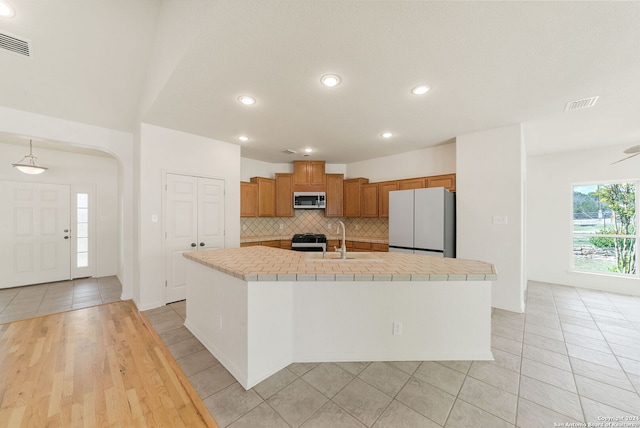 kitchen featuring tile countertops, a center island with sink, sink, tasteful backsplash, and appliances with stainless steel finishes