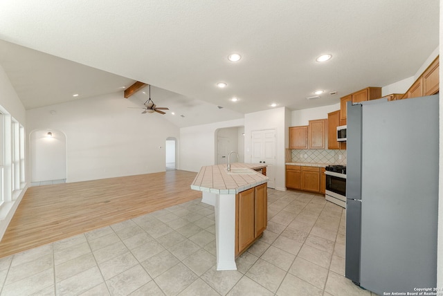 kitchen featuring vaulted ceiling with beams, gas range, stainless steel fridge, tile countertops, and a kitchen island with sink