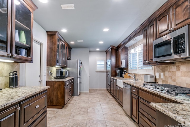 kitchen with appliances with stainless steel finishes, backsplash, dark brown cabinets, light stone countertops, and pendant lighting