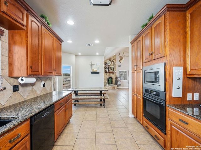 kitchen with dark stone counters, black appliances, light tile patterned floors, a stone fireplace, and backsplash