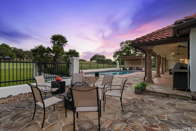 patio terrace at dusk featuring ceiling fan, a fenced in pool, and grilling area