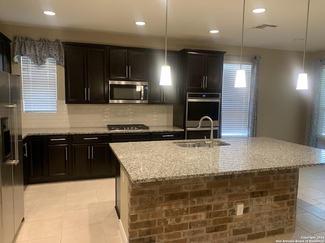 kitchen with sink, decorative light fixtures, appliances with stainless steel finishes, light stone countertops, and a kitchen island with sink