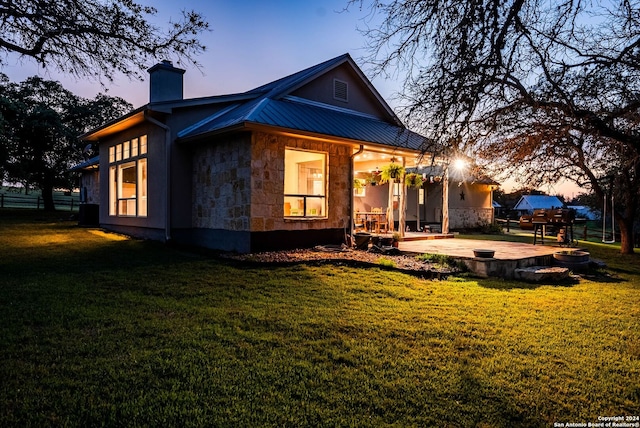 back of property at dusk featuring a patio, a chimney, a lawn, metal roof, and stone siding