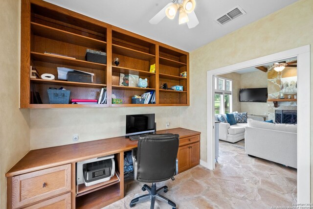 office with a stone fireplace, built in desk, and ceiling fan