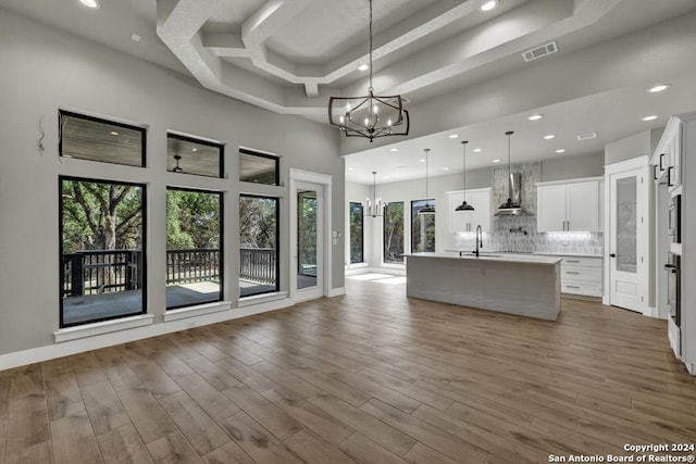 kitchen featuring an island with sink, a notable chandelier, pendant lighting, wall chimney range hood, and white cabinets