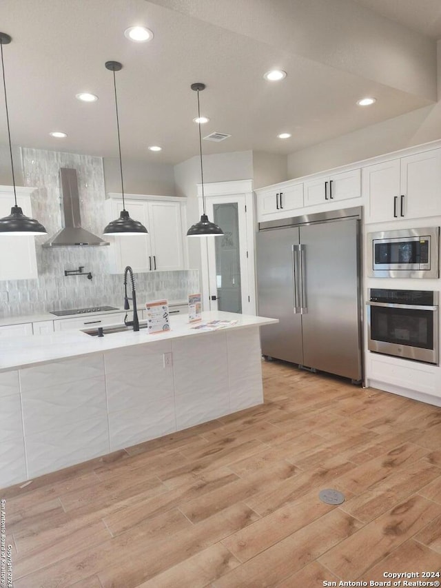 kitchen featuring decorative light fixtures, built in appliances, white cabinets, and wall chimney exhaust hood