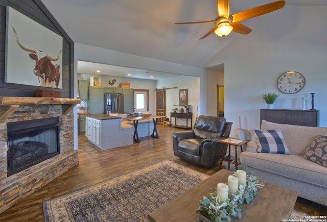 living room with ceiling fan, a barn door, vaulted ceiling, dark wood-type flooring, and a stone fireplace