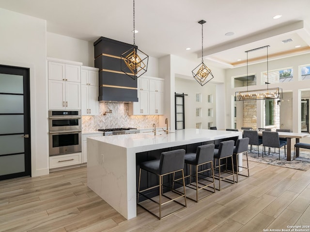 kitchen featuring a large island with sink, stainless steel double oven, hanging light fixtures, custom exhaust hood, and light hardwood / wood-style floors