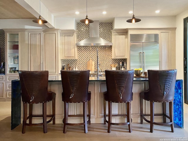 bar featuring pendant lighting, tasteful backsplash, built in refrigerator, cream cabinets, and wall chimney exhaust hood