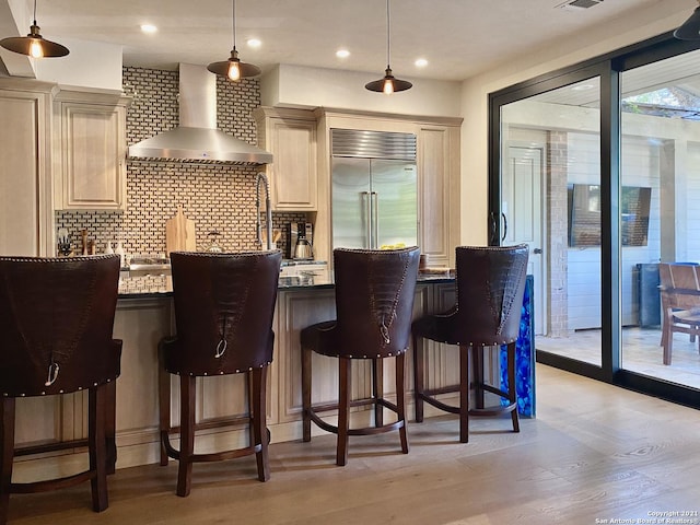 interior space featuring wall chimney range hood, backsplash, stainless steel built in refrigerator, decorative light fixtures, and light wood-type flooring