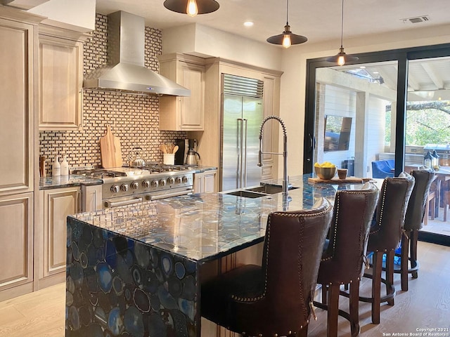 kitchen featuring wall chimney exhaust hood, premium appliances, an island with sink, dark stone counters, and backsplash