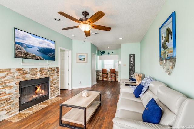 living room featuring ceiling fan, dark hardwood / wood-style flooring, a stone fireplace, and a textured ceiling