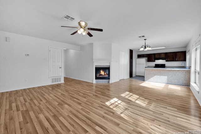 unfurnished living room featuring a tiled fireplace, sink, ceiling fan with notable chandelier, and light wood-type flooring