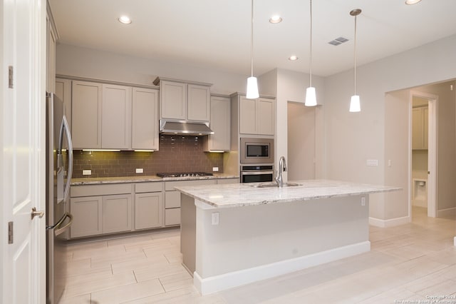 kitchen featuring tasteful backsplash, pendant lighting, an island with sink, light stone counters, and appliances with stainless steel finishes