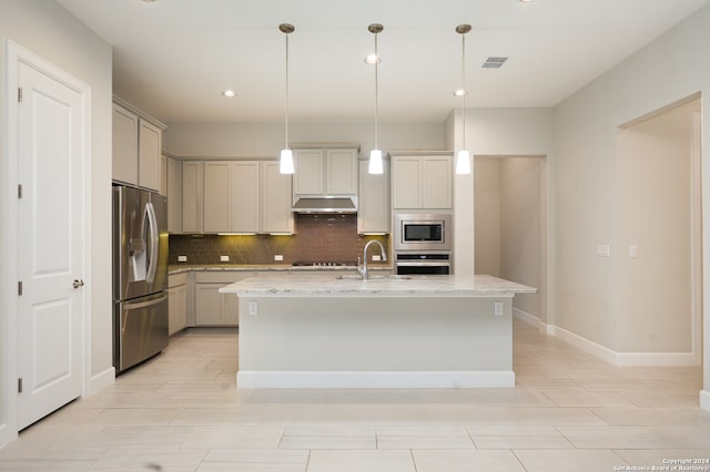 kitchen with appliances with stainless steel finishes, tasteful backsplash, a center island with sink, pendant lighting, and light stone counters