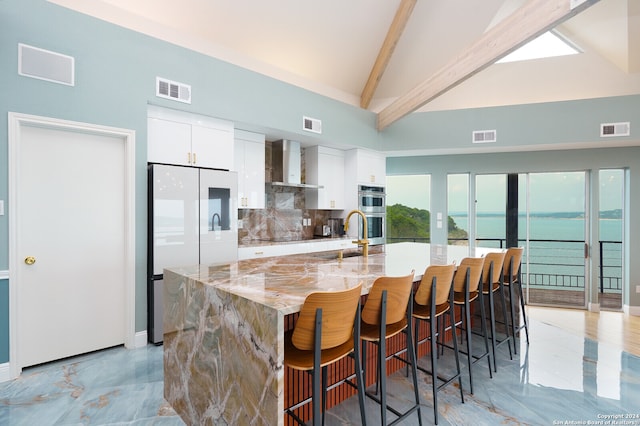 kitchen with high vaulted ceiling, white cabinetry, wall chimney exhaust hood, and an island with sink