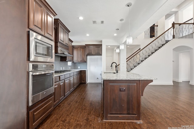 kitchen with a kitchen island with sink, dark hardwood / wood-style flooring, light stone countertops, hanging light fixtures, and appliances with stainless steel finishes