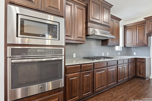 kitchen with light stone counters, ventilation hood, dark hardwood / wood-style flooring, appliances with stainless steel finishes, and backsplash