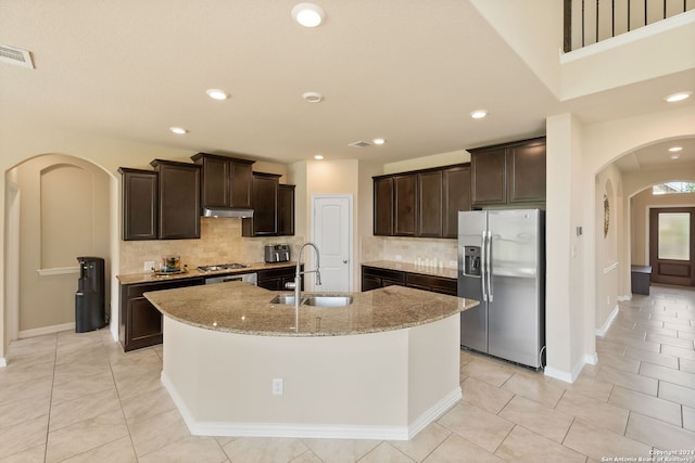 kitchen featuring an island with sink, sink, dark brown cabinetry, stainless steel appliances, and light stone countertops