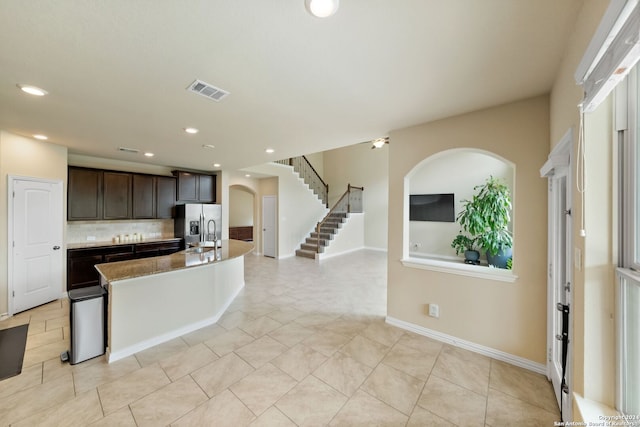 kitchen with light tile patterned flooring, backsplash, stainless steel fridge with ice dispenser, dark brown cabinets, and a center island with sink