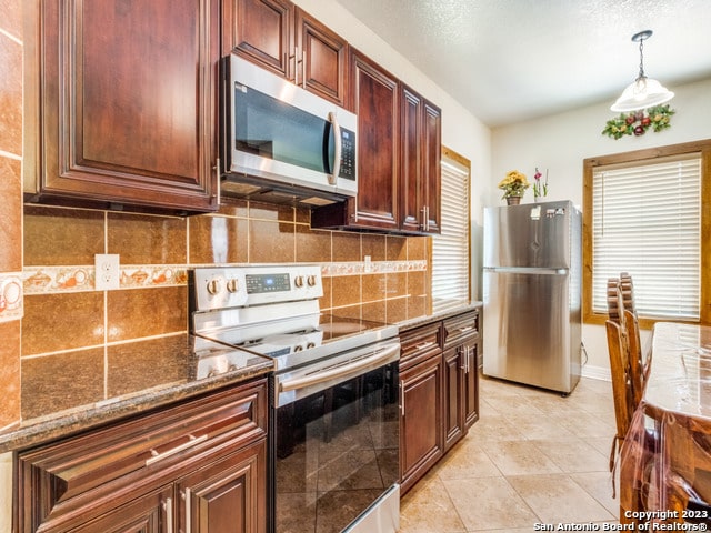 kitchen featuring appliances with stainless steel finishes, tasteful backsplash, hanging light fixtures, light tile patterned floors, and dark stone countertops