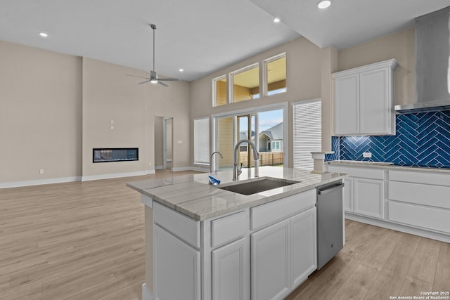 kitchen with a center island with sink, a sink, a glass covered fireplace, wall chimney exhaust hood, and dishwasher