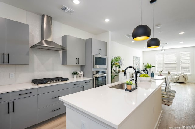 kitchen featuring appliances with stainless steel finishes, pendant lighting, an island with sink, sink, and wall chimney range hood