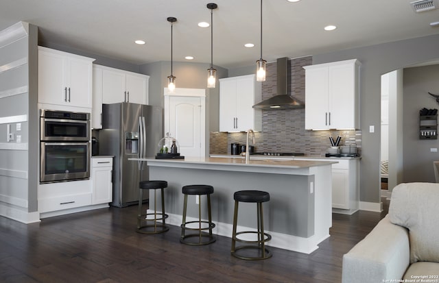 kitchen with appliances with stainless steel finishes, tasteful backsplash, a center island with sink, wall chimney range hood, and dark hardwood / wood-style floors