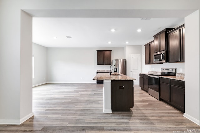 kitchen featuring stainless steel appliances, dark brown cabinetry, a kitchen island with sink, light wood-type flooring, and light stone countertops