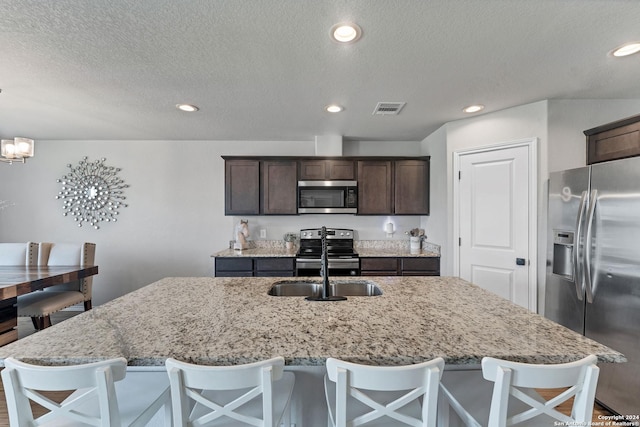 kitchen featuring sink, a kitchen island with sink, stainless steel appliances, light stone counters, and decorative light fixtures