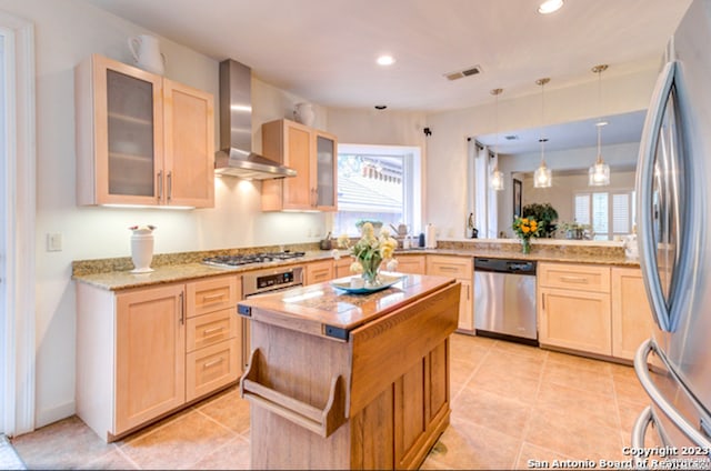 kitchen with appliances with stainless steel finishes, wall chimney exhaust hood, a kitchen island, and light brown cabinets