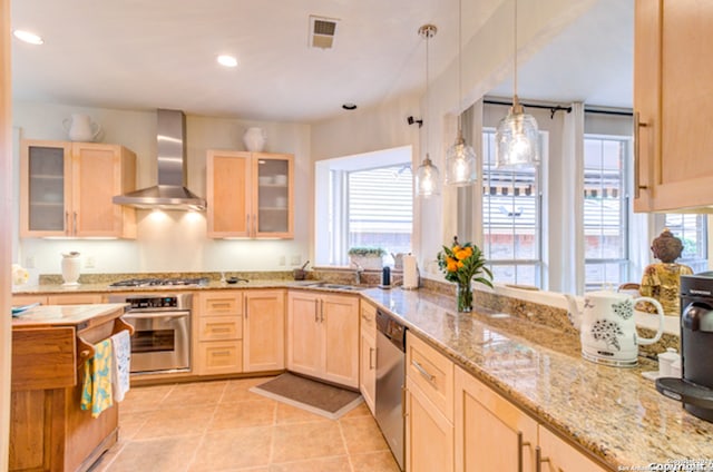 kitchen with appliances with stainless steel finishes, pendant lighting, light brown cabinets, light tile patterned floors, and wall chimney range hood