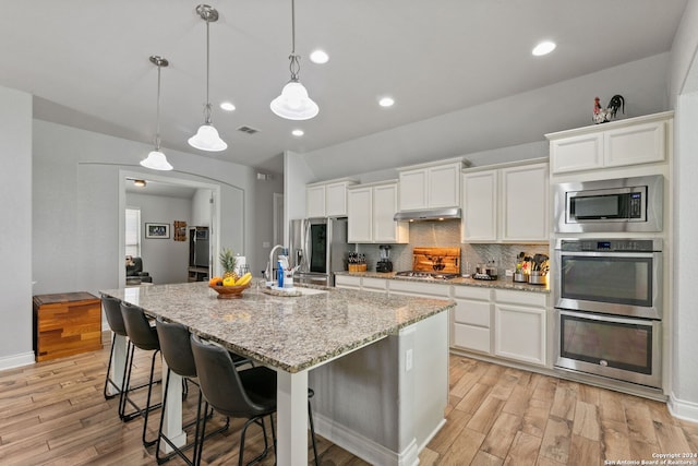 kitchen with appliances with stainless steel finishes, pendant lighting, tasteful backsplash, white cabinets, and a kitchen island with sink