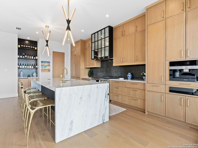 kitchen featuring wall oven, pendant lighting, an island with sink, sink, and light hardwood / wood-style flooring