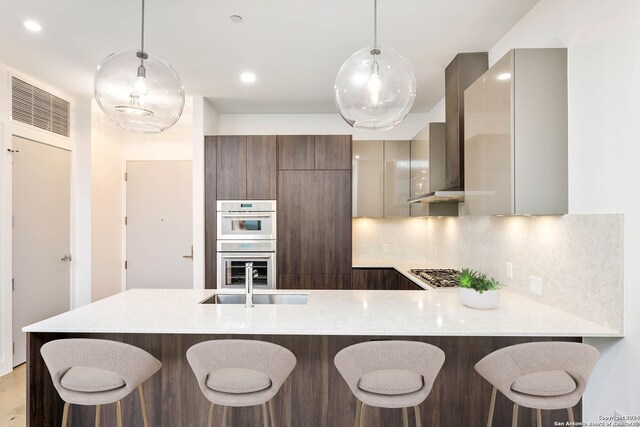 kitchen with white double oven, tasteful backsplash, wall chimney range hood, sink, and a kitchen breakfast bar