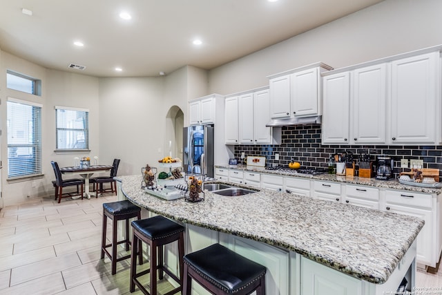 kitchen with white cabinets, backsplash, light stone counters, stainless steel appliances, and a center island with sink