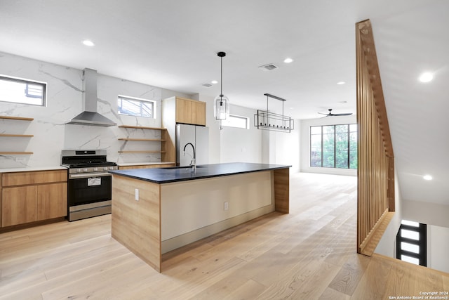 kitchen featuring wall chimney exhaust hood, a center island with sink, appliances with stainless steel finishes, light hardwood / wood-style floors, and decorative backsplash