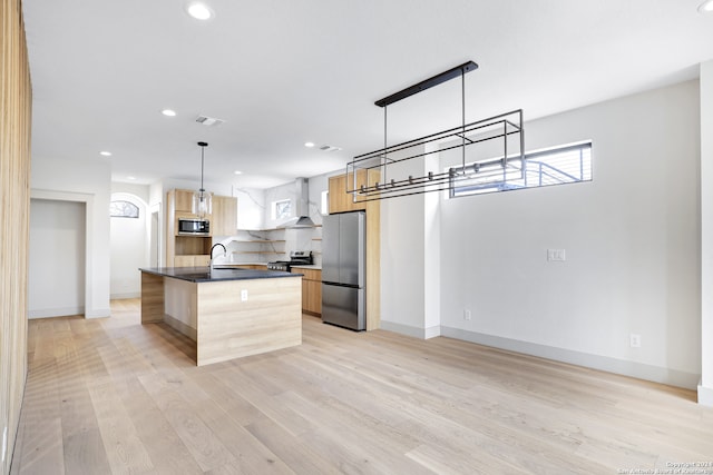 kitchen featuring wall chimney exhaust hood, light wood-type flooring, pendant lighting, stainless steel appliances, and a kitchen island with sink