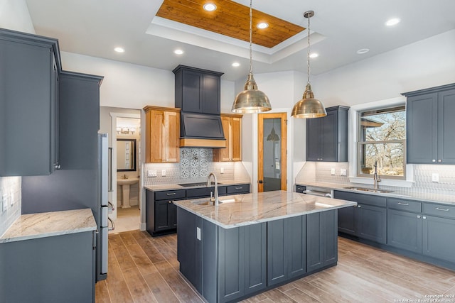 kitchen with pendant lighting, an island with sink, sink, a tray ceiling, and light stone countertops