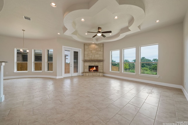 unfurnished living room featuring a tray ceiling, light tile patterned floors, a stone fireplace, and ceiling fan with notable chandelier