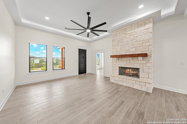 unfurnished living room featuring a stone fireplace, a tray ceiling, light wood-type flooring, and ceiling fan