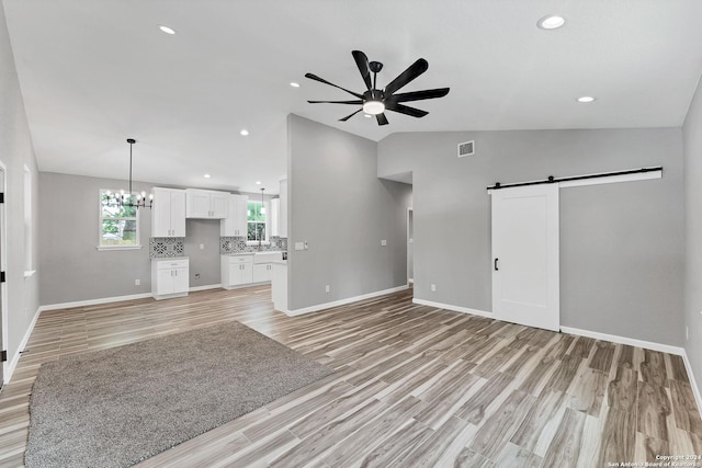 unfurnished living room featuring vaulted ceiling, a barn door, ceiling fan with notable chandelier, and light hardwood / wood-style floors