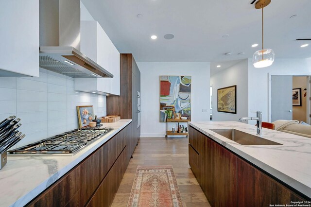 kitchen with wall chimney range hood, light hardwood / wood-style floors, hanging light fixtures, decorative backsplash, and sink