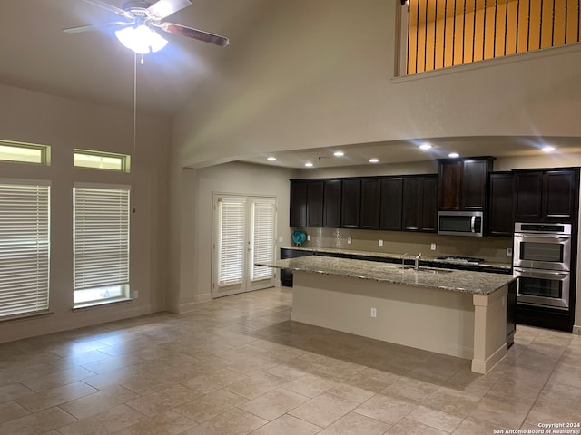 kitchen with appliances with stainless steel finishes, light stone counters, a kitchen island with sink, ceiling fan, and a towering ceiling