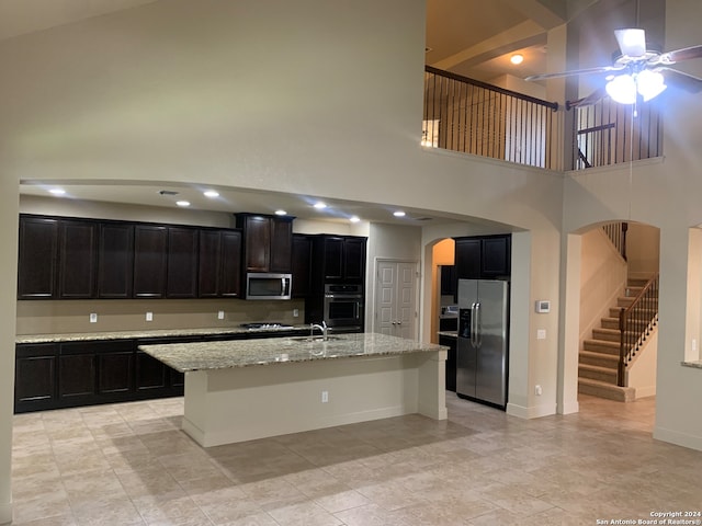 kitchen with ceiling fan, a center island with sink, appliances with stainless steel finishes, light tile patterned floors, and a towering ceiling