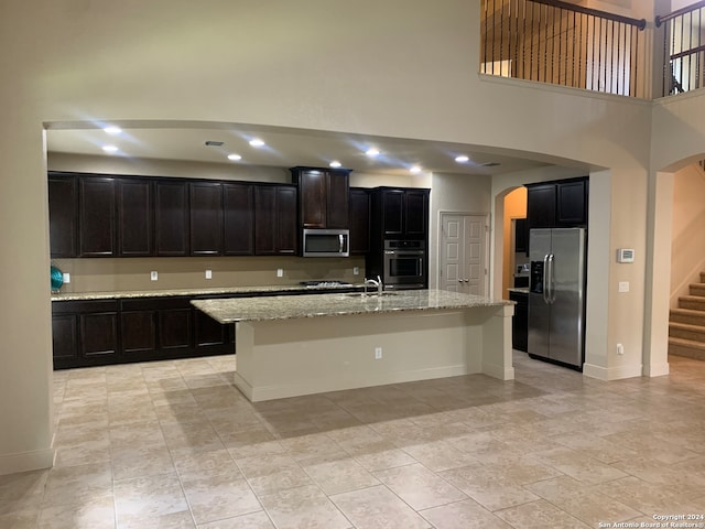 kitchen featuring light tile patterned flooring, a high ceiling, an island with sink, light stone counters, and appliances with stainless steel finishes