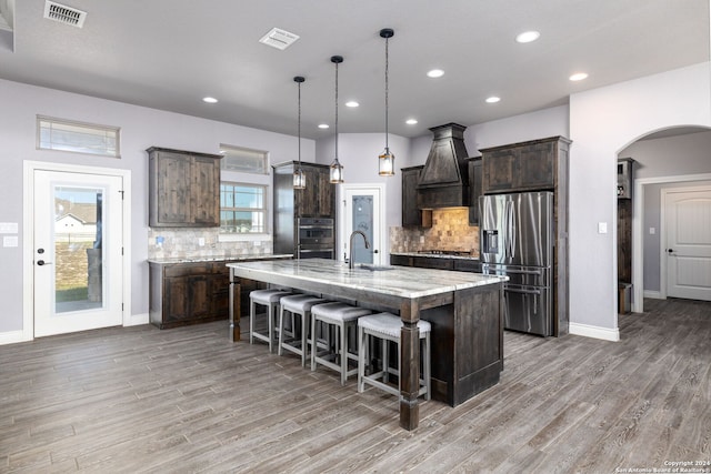 kitchen featuring dark brown cabinetry, light stone countertops, an island with sink, stainless steel fridge with ice dispenser, and decorative light fixtures