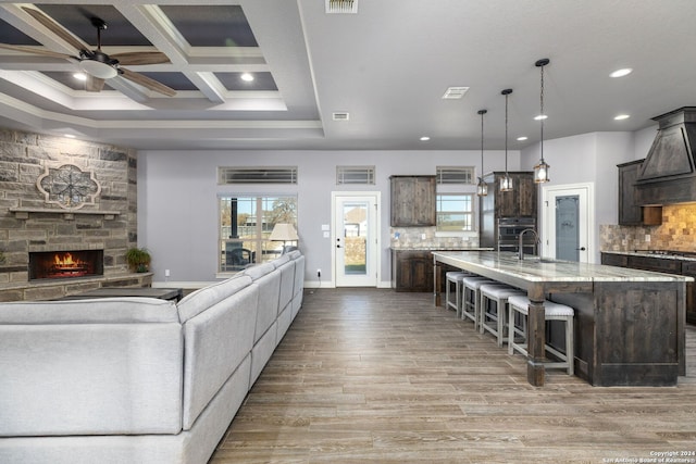 living room with coffered ceiling, a stone fireplace, wood-type flooring, beamed ceiling, and ceiling fan