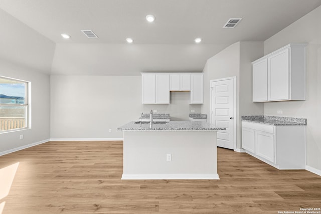 kitchen featuring white cabinetry, vaulted ceiling, an island with sink, light stone countertops, and light hardwood / wood-style floors